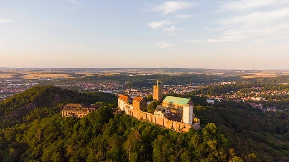 Blick von oben auf die Wartburg in Thüringen bei Eisenach, Burg steht erhöht auf einem Hügel und ist von viel grün umgeben, Burg wird von Abnedlicht erleuchtet, der Himmel ist balu