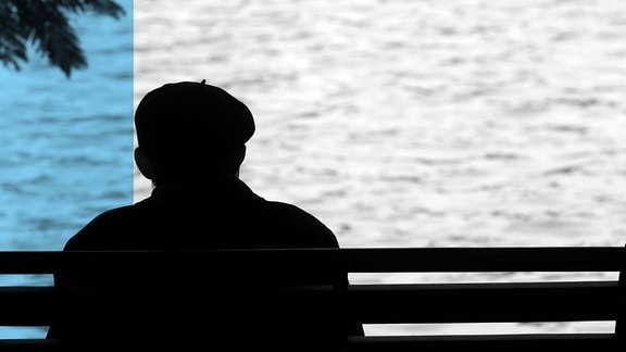 Silhouette eines Mannes, der vor einem Gewässer auf einer Bank sitzt.