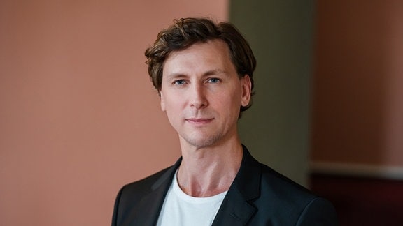 Rémy Fichet, neuer Ballettdirektor in Leipzig, ein Mann mit dunkelbloden Haaren, erträgt ein schwarzes Sakko über einem weißen Shirt. 