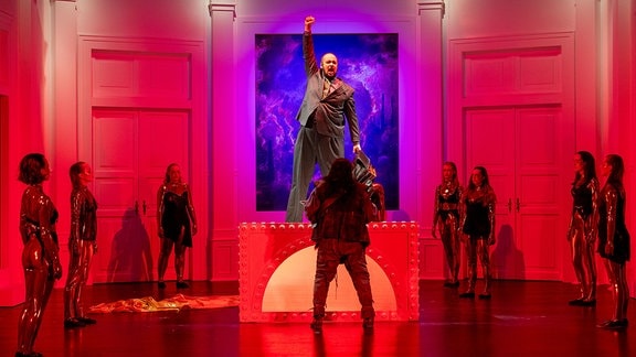 Die Bühne ist in rotes Licht getaucht: Eine Person steht auf einen Podest und streckt den Arm in die Höhe.
