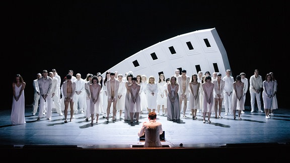 Eine Gruppe aus weiß gekleideten Menschen stehen auf einer Bühne vor einem Stück Mauer.