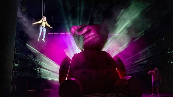 Ein riesiger Teddy auf einer Bühne wird von hinten angestrahlt und erzeugt ein Lichtspiel. Daneben hängt eine Person vom Bühnenhimmel.
