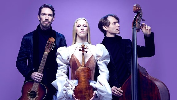 Eine Frau in einem weißen Kleid mit langen blonden Haaren hält eine Geige in den Händen, neben ihr stehen zwei schwar gekleidete Männer mit Streichinstrumenten. 