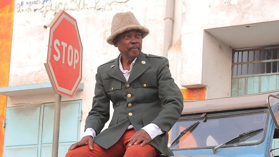 Musiker Roger Damawuzan mit Strohhut und roter Hose sitzt auf einem hellblauen Auto.