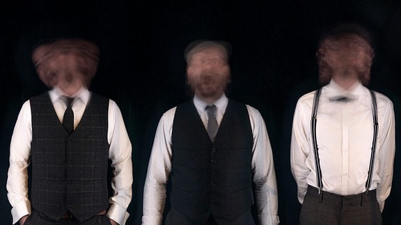 Die vier Mitglieder der Band Noon in weißen Hemden, Hostenträgern und schwarzen Westen stehen vor einem schwarzen Hintergrund. Ihre Gesichter sind verschwommen.