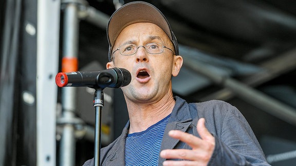 Steffen Mensching, ein Mann mit Brille udn Basecap, spricht in eine Mikrofon.