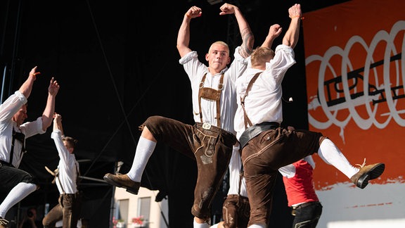 Tänzer in Lederhosen, weißen Hemden und weißen Strümpfen recken die Arme und tanzen im Kreis auf einer Bühne beim Rudolstadt-Festival 2023.