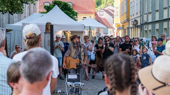 Straßenmusiker mit Hut und Gitarre in einer Fußgängerzone, im Kreis um ihn herum steht viel Publikum.