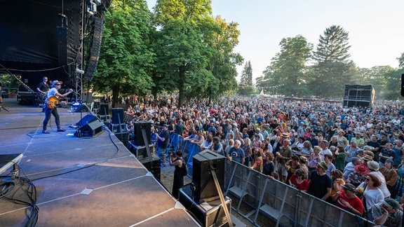 Dicht gedrängtes Publikum vor einer Open-Air-Bühne im Park beim Rudolstadt-Festival, ein Mann mit weißem Hut und Sonnenbrille greift nach dem Mikrofon.