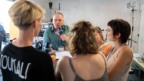 Ein Mann mit grünem Hemd und Brille hat ein Mikrofon in der Hand und spricht mit vier Frauen, die sich mit oihm an einem Tisch versammelt haben.