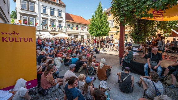 Eine Band spielt auf einer Bühne im Freien, die mit einem MDR KULTUR-Sonnensegel überdacht ist. Davor sitzen Zuhörerinnen und Zuhörer auf dem Boden oder stehen und hören zu.