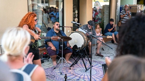 Eine Frau mit roten Harren spielt Gitarre, ein Mann singt neben einer Trommel, ein anderer spielt ebenfalls ein Instrument, drum herum stehen Leute in einer Fußgängerzone und hören zu.