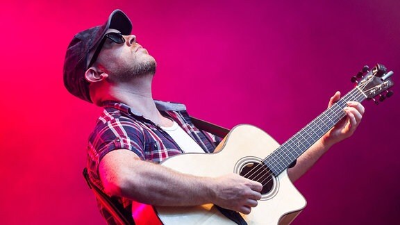 Ein Musiker steht mit Basecap und in kariertem Hemd auf der Bühne und spielt voller Hingabe Gitarre.