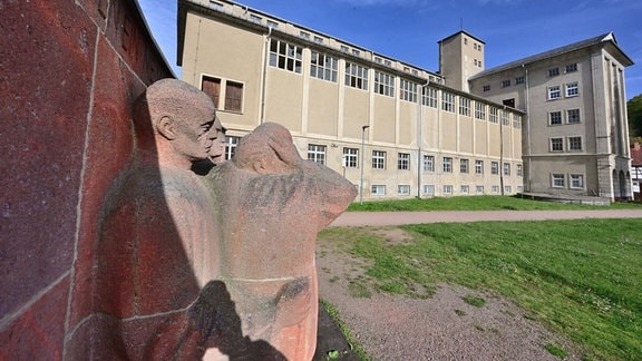 Blick vorbei an einem Figurenrelief auf ein schlichtes Gebäude.