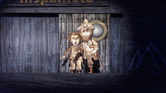 Zwei hellbraune Puppen eines Kapitäns und eines Kindes stehen vor einer hölzernen Wand.