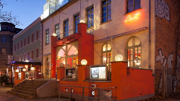 Außenansicht der Schaubühne Lindenfels Leipzig: ein zweistöckiges beleuchtetes Gebäude mit Terrasse und markantem Eingang in abendliches Licht getaucht 
