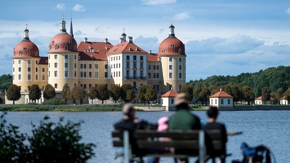 Drei Personen sitzen auf einer Bank an einem Fluss, im Hintergrund ist Schloss Moritzburg zu sehen. 