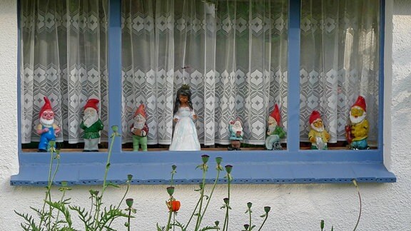 Schneewittchenfigur und sieben Gartenzwerge aufgereiht im Fenster eines Gartenhauses