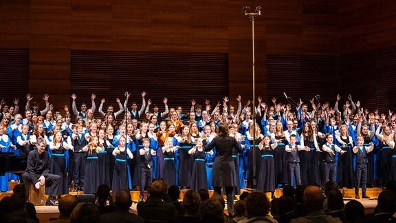 Ein großer Chor in mit blauen Tüchern steht auf einer holzvertäfelten Bühne.