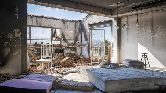 Blick in eine verwüstete Wohnung: Die Wände sind angekokelt, zwischen Unrat liegen zwei Matratzen und stehen Stühle. Durch das fehlende Dach ist blauer Himmel zu sehen.