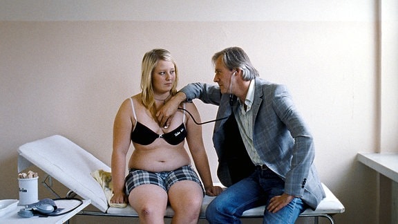 Eine junge Frau mit BH und Shorts bekleidet sitzt auf einer Arztliege, daneben ein älterer Mann, der sie abhört.