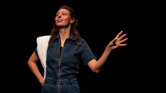 Eine Frau in einem Jeans-Kleid steht auf der Bühne und gestikuliert mit ihrer linken Hand.