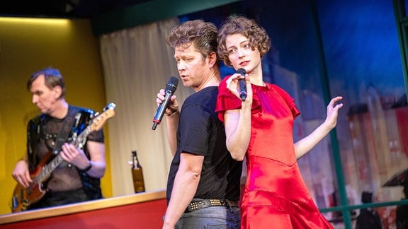 Ein Mann in T-Shirt und eine Frau in rotem Kleid singen Rücken und Rücken in Mikros.