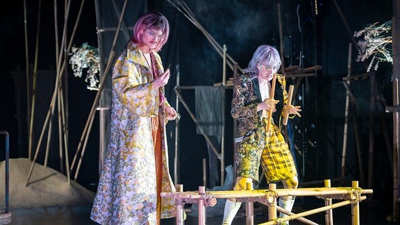 Zwei Menschen mit bunten Haaren und greller Kleidung stehen vor einer Holzkonstruktion auf einer Bühne.