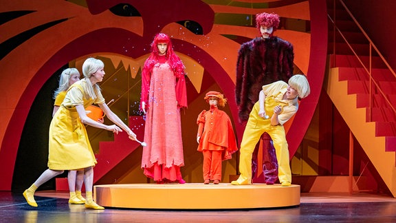 Drei Personen in gelber Kleidung bewegen sich auf einer orangefarbenen Bühne um drei Puppen in rötlicher Kleidung.