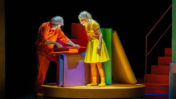 Eine Frau in einem gelben Kleid und ein Mann in einem orangenen Overall schauen auf einen leuchtenden Bildschirm.