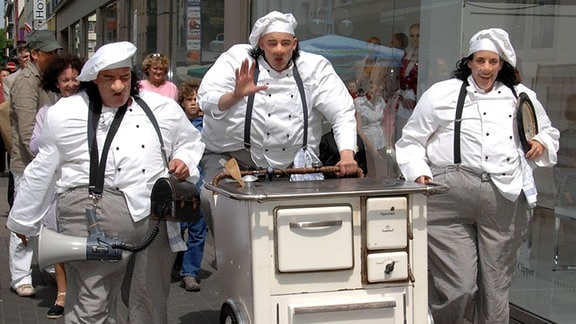 Drei als dicke Bäcker verkleidete Schauspieler schieben einen Crêpe-Stand durch die Straße.