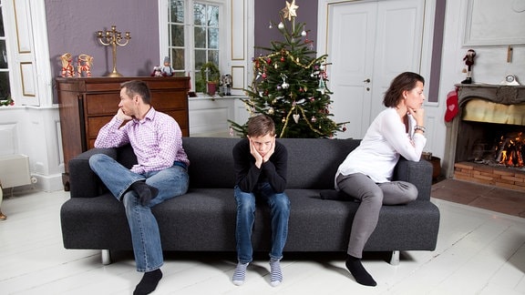 Mann Kind und Frau sitzen, voneinander abgewandt auf einem Sofa, vor einem Weihnachtsbaum