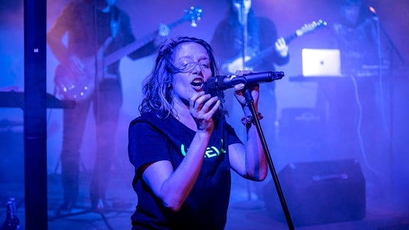 Eine Person singt gefühlvoll in eine Mikro, im Hintergrund spielt eine Band.