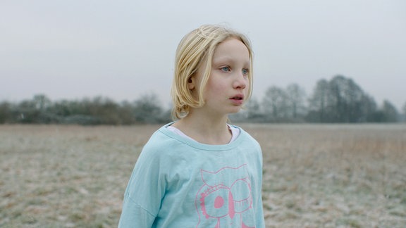 Filmstill, ein blondes Mädchen steht auf einem Feld.