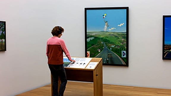 Ein Mann fingert an einer Medienstation auf einem holzbrauenen Tisch vor einem Gemälde, das eine Straße durch grüße Hügel vor einem blauen Horizont zeigt.