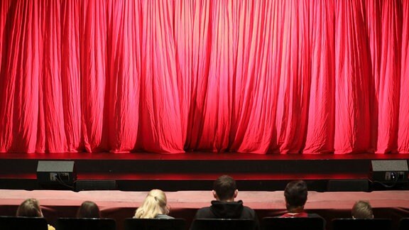 Besucher sitzen vor einem roten Theatervorhang.