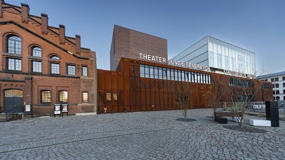 Blick auf einen gepflasterten Hof, auf dem ein altes und ein modernes Gebäude nebeneinander stehen. 