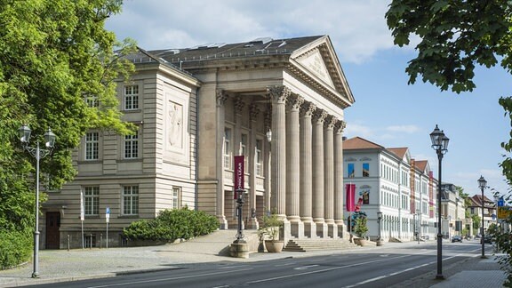 Zu sehen ist das Theater Meiningen von der Seite bei blauem Himmel, vor dem Gebäude mit Spitzdach und Säulen liegt direkt die Straße, links steht ein Baum