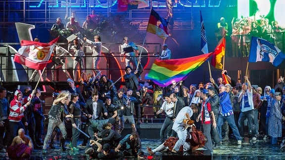 Bühne mit voelen Menschen, manche schwenken Fahnen, darunter eine Regenbogenfahne und Banner von Fußballvereinen