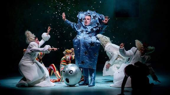 Szene aus Oper "Alice im Wunderland", ein Mann in einem blauen Kostüm, zu seinen Füßen mehrere Personen in weißen Kostümen.