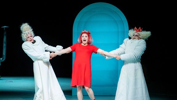 Eine Frau in einem roten Kleid mit roten Haaren steht auf einer Bühne, zwei Personen in weißen Kostümen ziehen an ihren Armen.  