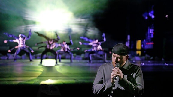 Ein Mann mit schwarzer Mütze steht vor einem Mikro. Im Hintergrund tanzen Menschen in schwarzen Hosen.