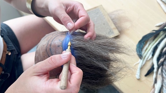 Ein Bart wird von einer Maskenbildnerin an einen Holzkopf geknüpft.
