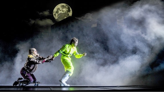 Zu sehen ist eine Szene aus "Das Schloss" am Staatsschauspiel Dresden: Ein Mann in neongelben Warn-Klamotten und Regenstiefeln zieht eine Person auf einem Skateboard über die Bühne.