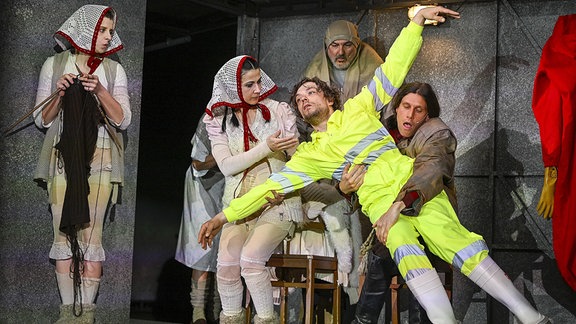 Zu sehen ist eine Szene aus "Das Schloss" am Staatsschauspiel Dresden: Ein Mann in neongelben Warn-Klamotten streckt alle Gliedmaßen von sich und wird von einem anderen Mann gehalten, neben ihnen stehen zwei Frauen, eine davon strickt