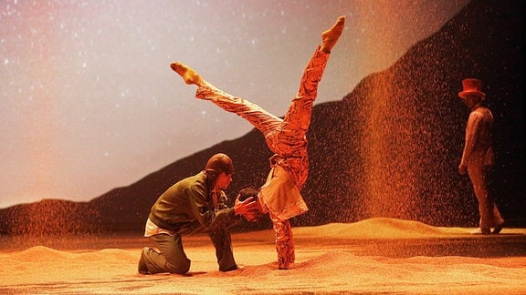 Zu sehen ist eine Szene aus "Der kleine Prinz": zwei Figuren befinden sich in einer Wüste, eine macht einen Handstand mit gespreizten Beinen, die andere kniet davor und hält den Kopf