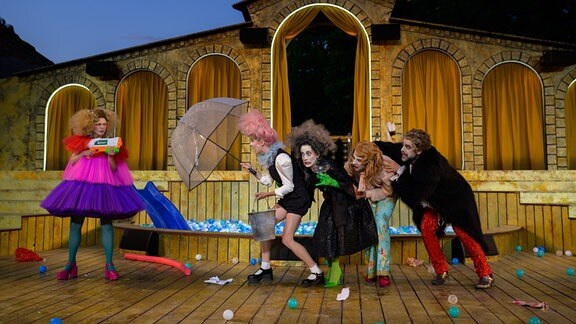 Vier Personen verstecken sich hinter einem Schirm vor einer Frau. Alle sind schrill kostümiert und geschminkt.