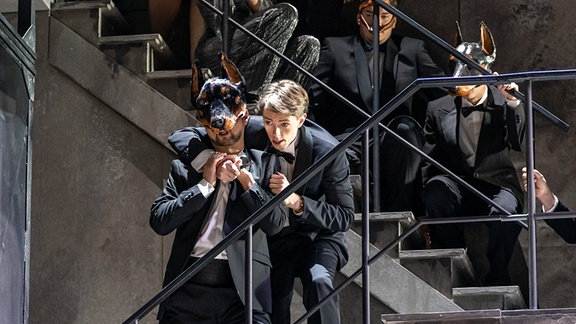 Don Giovanni steht mit seinen Partygästen auf einer Treppe, die meisten tragen Anzüge und Dobermann-Masken