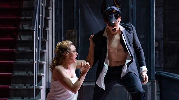 Donna Anna greift im Handtuch bekleidet an die Schulter von Don Giovanni, der eine Dobermann-Maske trägt und schreit