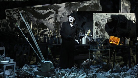 Ein Mann in schwarzem Anzug und Hut steht in einem chaotischen Atelier, der Boden ist voller zerknüllter Zeitungen.